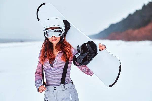 Portrait d'une belle rousse en casque protecteur et lunettes tenant un snowboard, posant sur une plage enneigée — Photo
