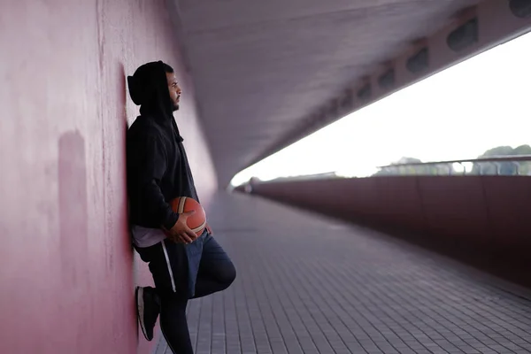 Afro-Amerikaanse man, het dragen van sportkleding houden een basketbal terwijl leunend op een muur buiten — Stockfoto