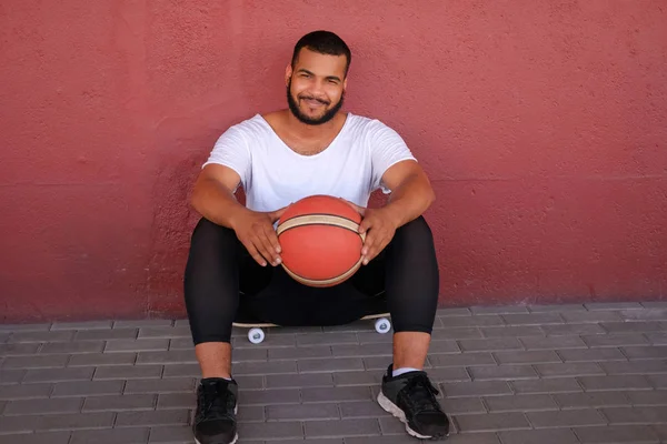 Afroamerikansk man bära sportkläder sitter på en skateboard och hålla en basket medan lutande på en vägg utanför — Stockfoto