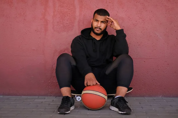 Afro-americano vestindo um capuz preto sentado em um skate e segurando um basquete enquanto se inclina em uma parede do lado de fora — Fotografia de Stock