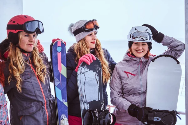 Vista lateral Retrato de una mujer amiga en ropa deportiva de invierno sosteniendo tablas de snowboard y esquís posando juntas cerca de la costa nevada del mar —  Fotos de Stock