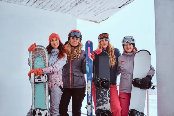 Vista lateral Retrato de uma mulher amigas em esportes roupas de inverno segurando snowboards e esquis posando juntas perto da costa do mar nevado — Fotografia de Stock