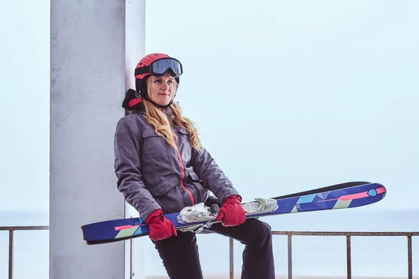 Mujer rubia con casco protector y gafas que sostienen esquís mientras está sentada en una barandilla cerca de la costa nevada del mar —  Fotos de Stock