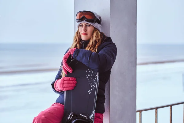 Mujer rubia en sombrero de invierno y gafas que sostienen una tabla de snowboard mientras está sentado en una barandilla cerca de la costa del mar nevado —  Fotos de Stock