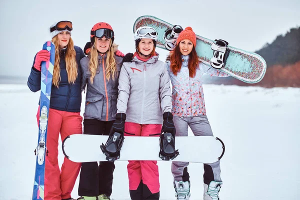 Přítelkyně v sportovní zimní oblečení, snowboardy a lyže stáli spolu v objetí a při pohledu na fotoaparát — Stock fotografie