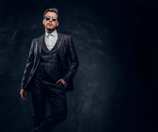 Ein stilvoller junger Mann im eleganten Anzug mit Sonnenbrille posiert mit einer Hand in der Tasche vor einer dunklen, strukturierten Wand — Stockfoto