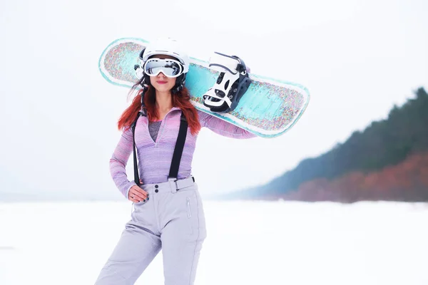 Porträt eines schönen rothaarigen Mädchens mit Schutzhelm und Schutzbrille, das ein Snowboard hält und an einem verschneiten Strand posiert — Stockfoto