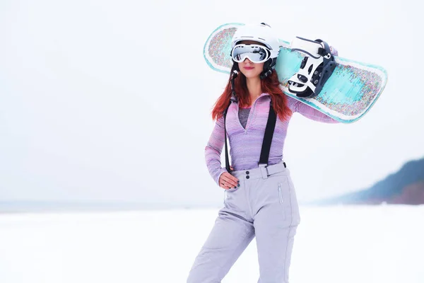 Porträt eines schönen rothaarigen Mädchens mit Schutzhelm und Schutzbrille, das ein Snowboard hält und an einem verschneiten Strand posiert — Stockfoto