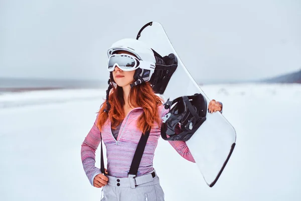 Porträt eines schönen rothaarigen Mädchens mit Schutzhelm und Schutzbrille, das ein Snowboard hält und an einem verschneiten Strand posiert — Stockfoto