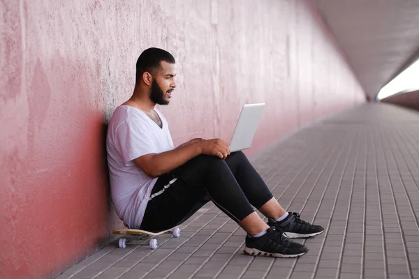 African-American guy is sitting on a bridge sidewalk, holding the laptop on his knees and chatting with friends — Stock Photo, Image