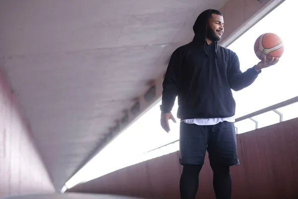 Afrika-Amerika pria mengenakan hoodie hitam memegang bola basket sambil berdiri di jembatan trotoar — Stok Foto