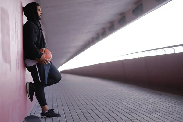 Ragazzo afro-americano indossa abbigliamento sportivo in possesso di una pallacanestro mentre si appoggia su un muro esterno — Foto Stock