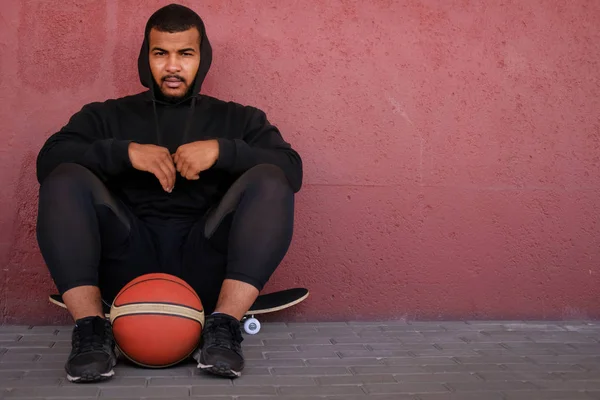 Orang Afrika-Amerika mengenakan hoodie hitam duduk di skateboard dan memegang bola basket sambil bersandar di dinding luar — Stok Foto