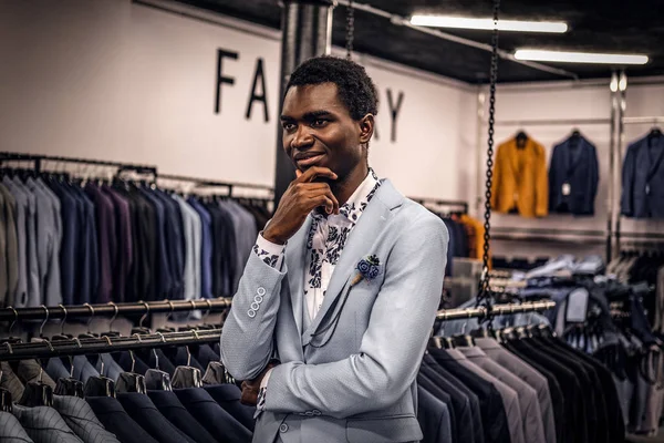 Un hombre africano elegantemente vestido posando con la mano en la barbilla mientras está de pie en una tienda de ropa masculina clásica . — Foto de Stock