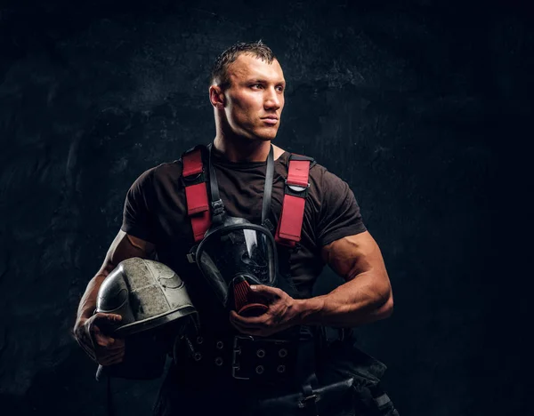 Bombero musculoso guapo sosteniendo un casco y una máscara de oxígeno de pie en el estudio contra una pared de textura oscura — Foto de Stock