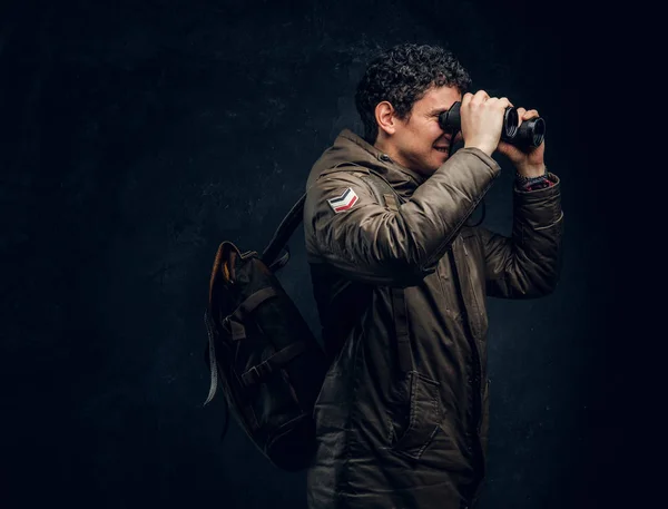 Viajante em uma roupa de caminhada com uma mochila olha através de binóculos no estúdio contra uma parede escura — Fotografia de Stock