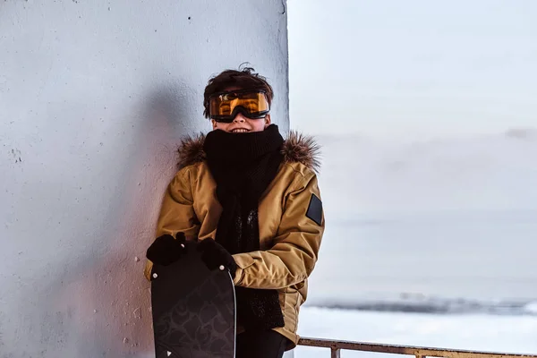 Ένας νεαρός snowboarder ντυμένος με ζεστά ρούχα και προστατευτικά γυαλιά, κάθεται στο κιγκλίδωμα κοντά στην παραλία χιονισμένο — Φωτογραφία Αρχείου