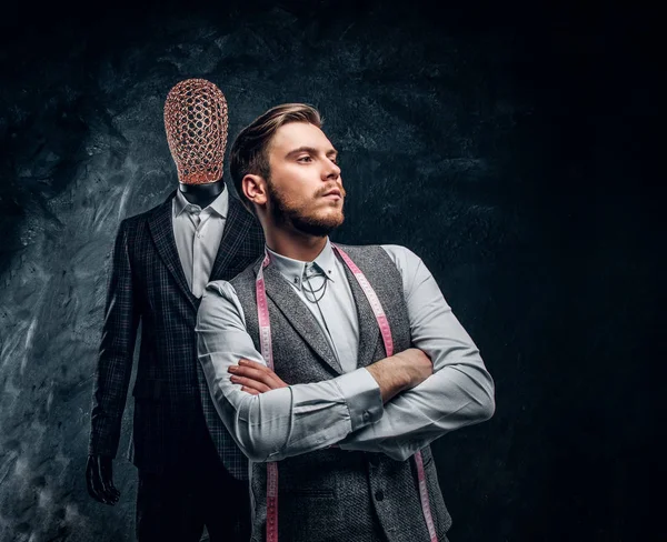 Un joven diseñador de moda confiado posando junto a un maniquí en traje exclusivo hecho a medida para hombre en un estudio de sastre oscuro — Foto de Stock