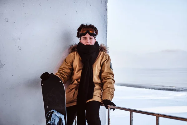 Um jovem snowboarder vestido com roupas quentes e óculos de proteção sentado em trilhos perto da praia nevada — Fotografia de Stock