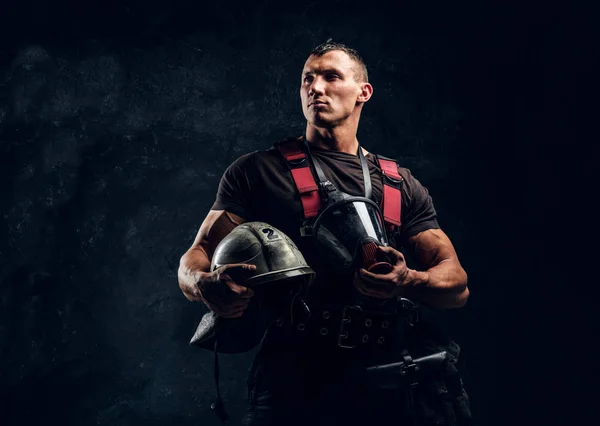 Handsome muscular fireman holding a helmet and oxygen mask standing in the studio against a dark textured wall — Stock Photo, Image