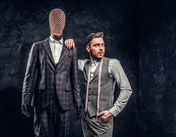 Un joven diseñador de moda confiado posando junto a un maniquí en traje exclusivo hecho a medida para hombre en un estudio de sastre oscuro —  Fotos de Stock