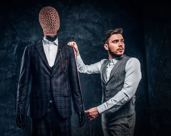 Un diseñador de moda con cinta métrica comprueba la longitud de las mangas de un elegante traje de hombre hecho a medida en un estudio de sastre oscuro — Foto de Stock