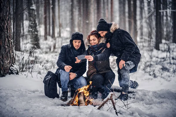 Grupo de amigos alegres viajantes fazer Sefley sentado ao lado da fogueira no meio de uma floresta nevada — Fotografia de Stock