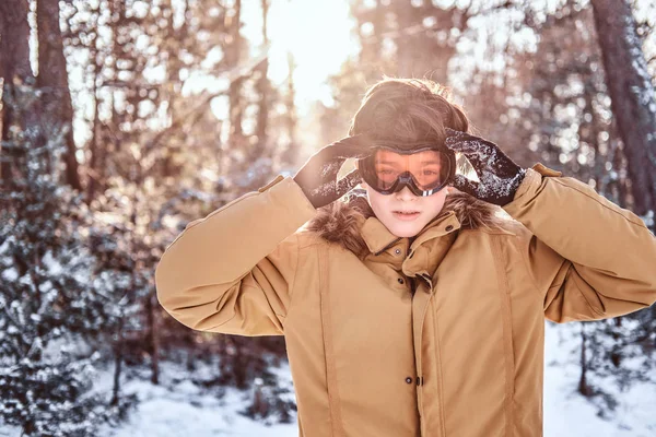 Retrato de um jovem snowboarder vestido com roupas quentes e óculos de proteção em pé em uma floresta nevada durante o pôr do sol — Fotografia de Stock