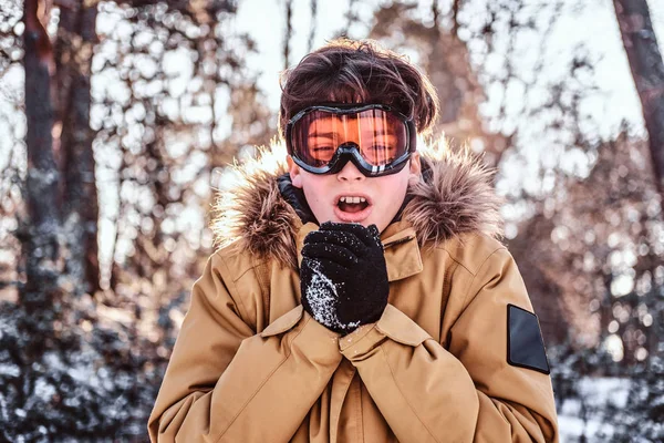 젊은 snowboarder 따뜻한 옷과 고글, 옷을 입고 눈 덮인 숲에 서 있는 그의 손을 따뜻하게합니다 — 스톡 사진