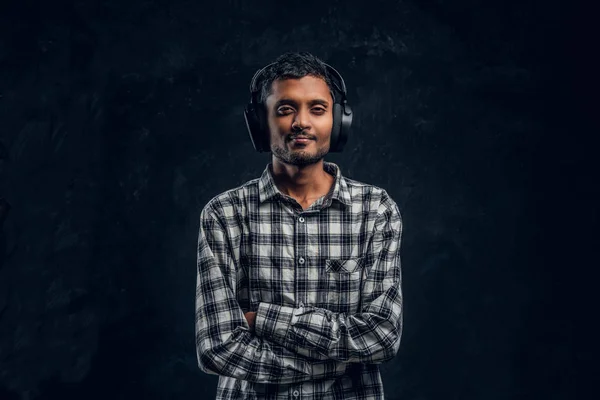 Knappe Indiase jongen is gelukkig luisteren naar muziek in draadloze hoofdtelefoons permanent met zijn armen gekruist in een donkere studio — Stockfoto