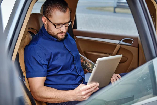 Homem de negócios barbudo sentado em um banco de trás no carro de luxo, trabalhando com um tablet — Fotografia de Stock