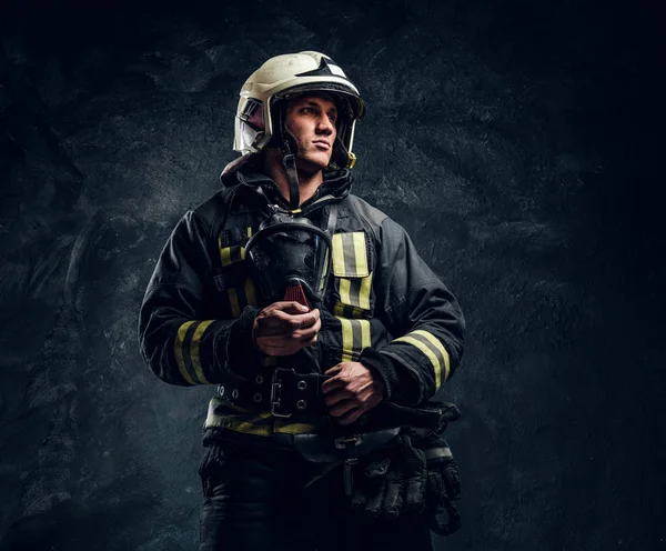 Bombero brutal en uniforme y casco de seguridad sosteniendo una máscara de oxígeno y mirando hacia los lados con una mirada segura . — Foto de Stock