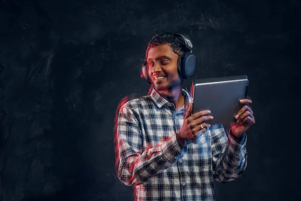 Chico indio en auriculares inalámbricos sosteniendo una tableta y disfruta escuchando su música favorita, sonriendo y mirando hacia otro lado . — Foto de Stock