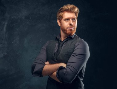 Elegantly dressed redhead man posing with his arms crossed in studio against a dark textured wall clipart