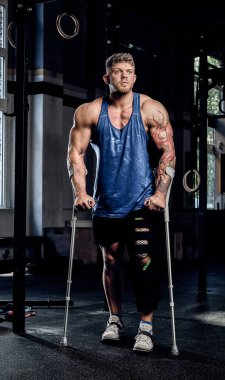 Kaslı vücut geliştirmeci koltuk değneği ve bir bacak bir spor salonunda bandaj ile ayakta.