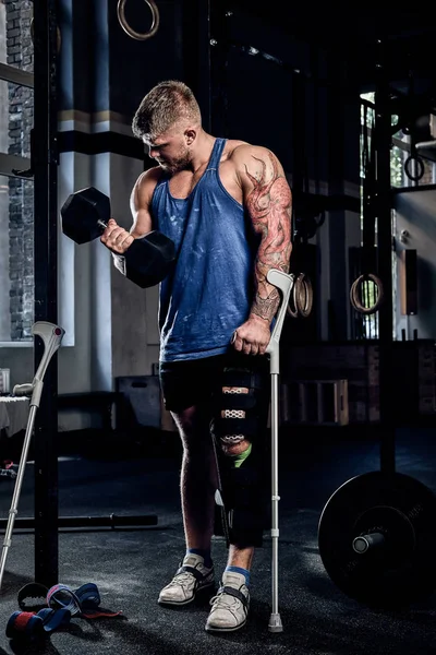 Professional bodybuilder with a tattoo on his hand, warming up on a gym, doing exercises with a dumbbell while leaning on a crutch. — Stock Photo, Image