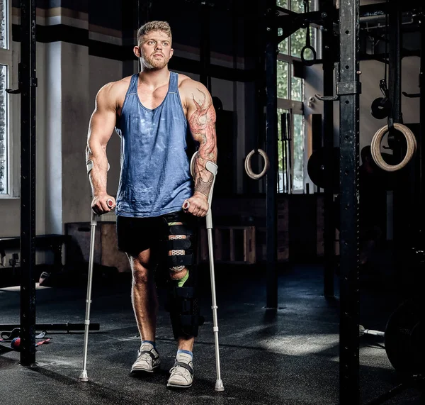 Muscular bodybuilder standing with crutches and bandage on a leg in a gym.