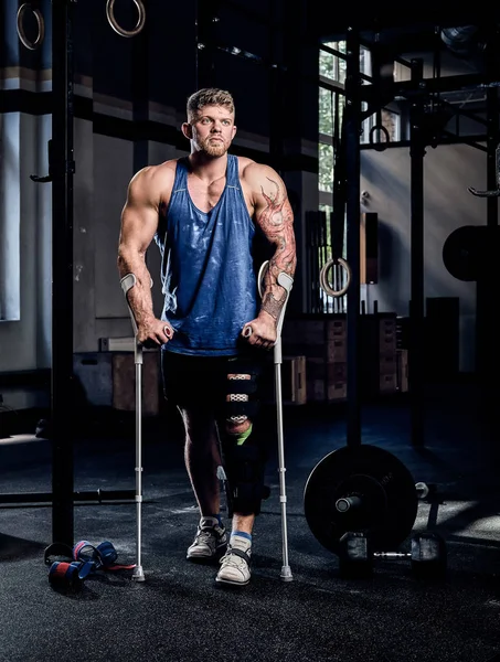 Gespierde bodybuilder staande met krukken en pleister op een been in een sportschool. — Stockfoto