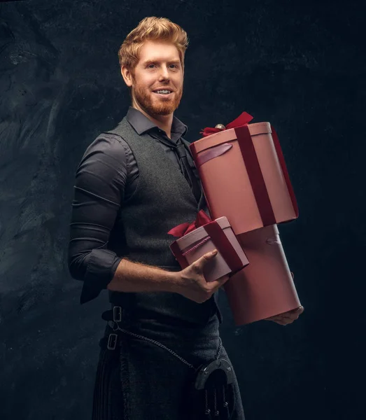 Redhead man wearing kilt and sporran holding gifts while posing in a dark studio