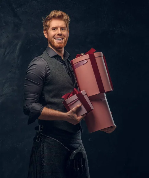 Redhead man wearing kilt and sporran holding gifts while posing in a dark studio
