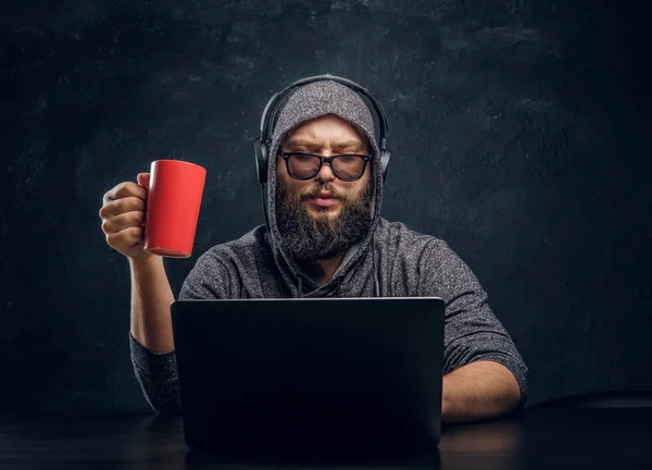 Um hacker senta-se atrás de um laptop, segurando uma xícara de café e trabalhando com programas em uma sala escura Imagens Royalty-Free