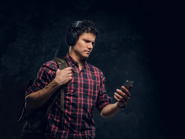 Hombre joven en auriculares inalámbricos escucha música y mantiene el teléfono inteligente en un estudio contra el fondo de la pared oscura — Foto de Stock