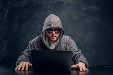 Programcı hoodie ve karanlık bir ofiste bir masada oturan dizüstü bilgisayarda parola kesmek güneş gözlüğü