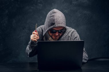Bir bıçak kilit-up bir dizüstü karanlık bir ofiste arkasında oturan hoodie ve güneş gözlüğü hacker tutar