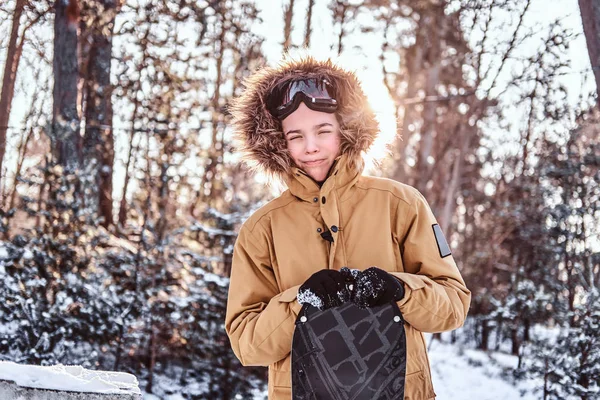 Jeune snowboardeur vêtu de vêtements chauds s'appuie sur un snowboard debout dans une forêt enneigée au coucher du soleil — Photo