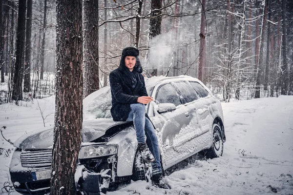 Bilen hamnade i en skid och kraschade in i ett träd på en snöig väg. En föraren sitter på motorhuven och röker en cigarett i väntan på en bärgningsbil — Stockfoto