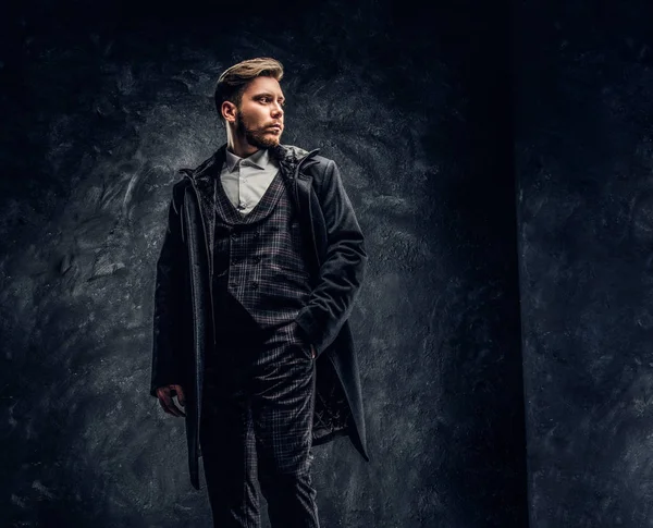 Um homem elegante vestido com um terno elegante e casaco posando com uma mão em um bolso contra uma parede de textura escura — Fotografia de Stock