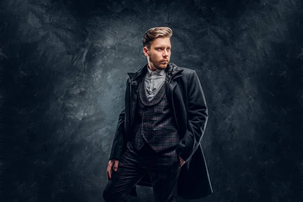 En snygg man klädd i en elegant kostym och kappa som poserar med en hand i fickan mot en mörk texturerat vägg — Stockfoto