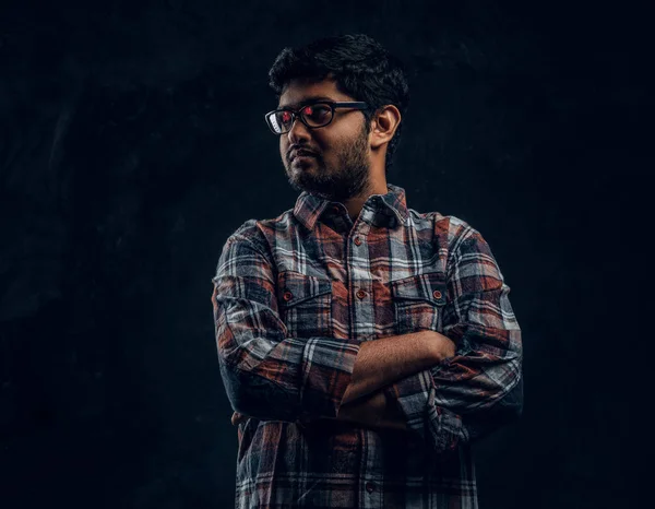 一个年轻的印度小伙子穿着眼镜和格子衬衫的工作室肖像, 他的双臂交叉, 侧身看 — 图库照片