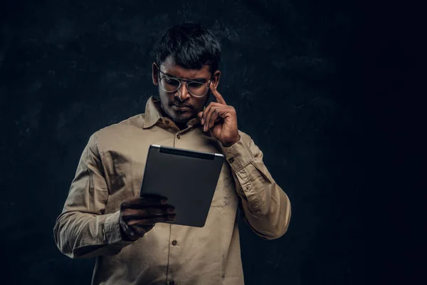 Hombre indio pensativo en gafas y camisa usando una tableta en el estudio —  Fotos de Stock